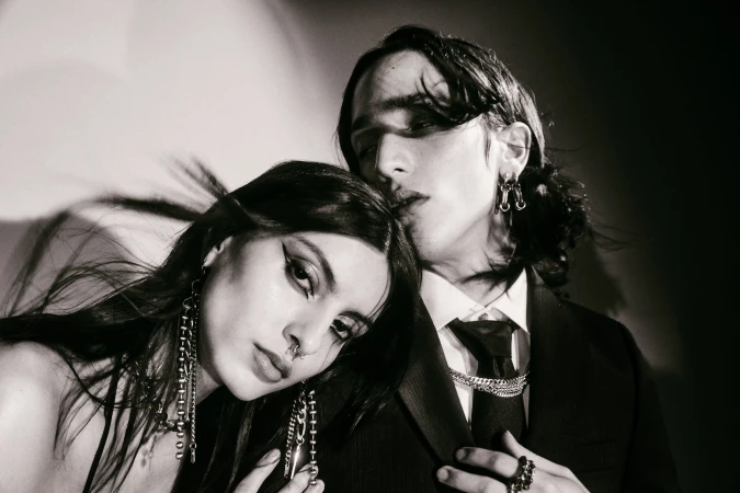 Monochrome portrait of a fashionable couple, a woman with dramatic eye makeup and a man in a suit, posing in a studio setting.