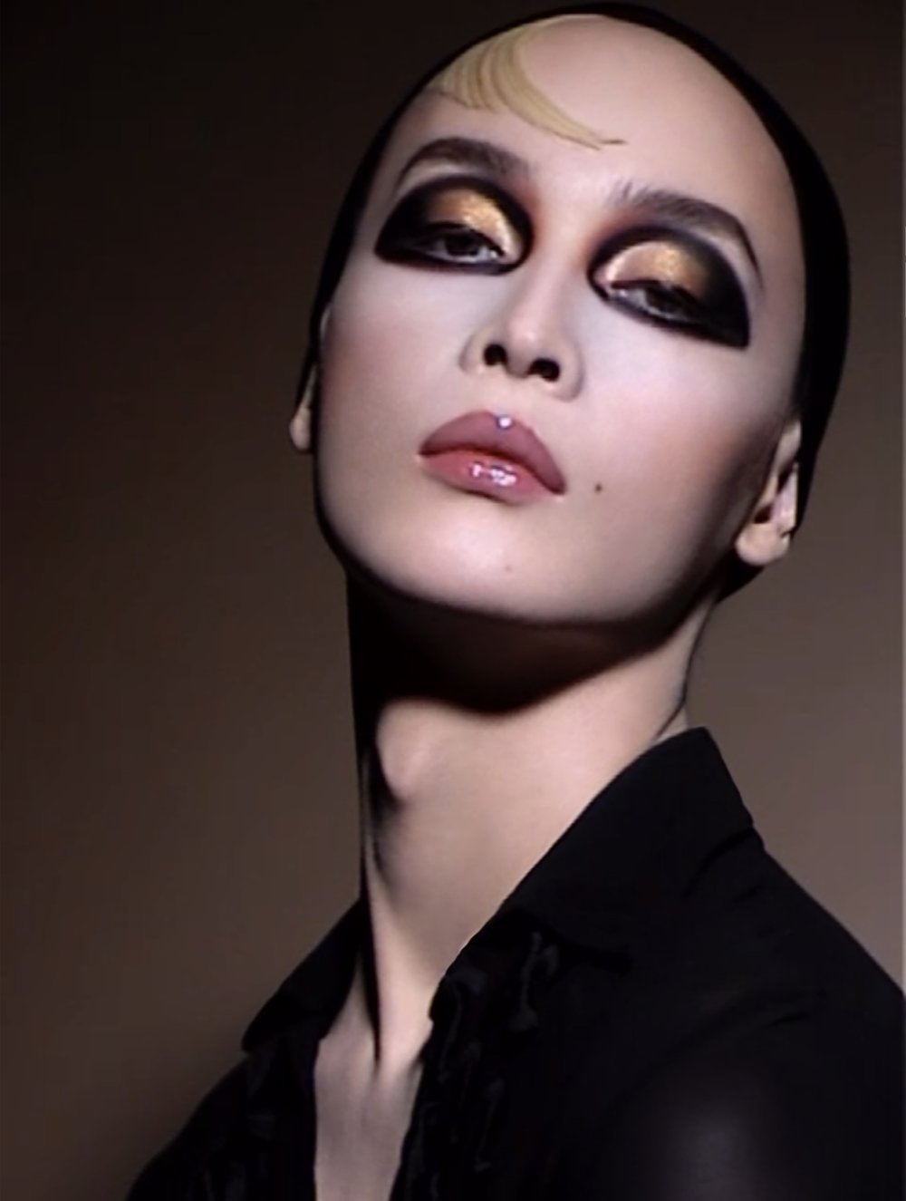 Close-up portrait of a person with dramatic gold and black eye makeup, glossy lips, and a dark shirt, set against a dark backdrop.