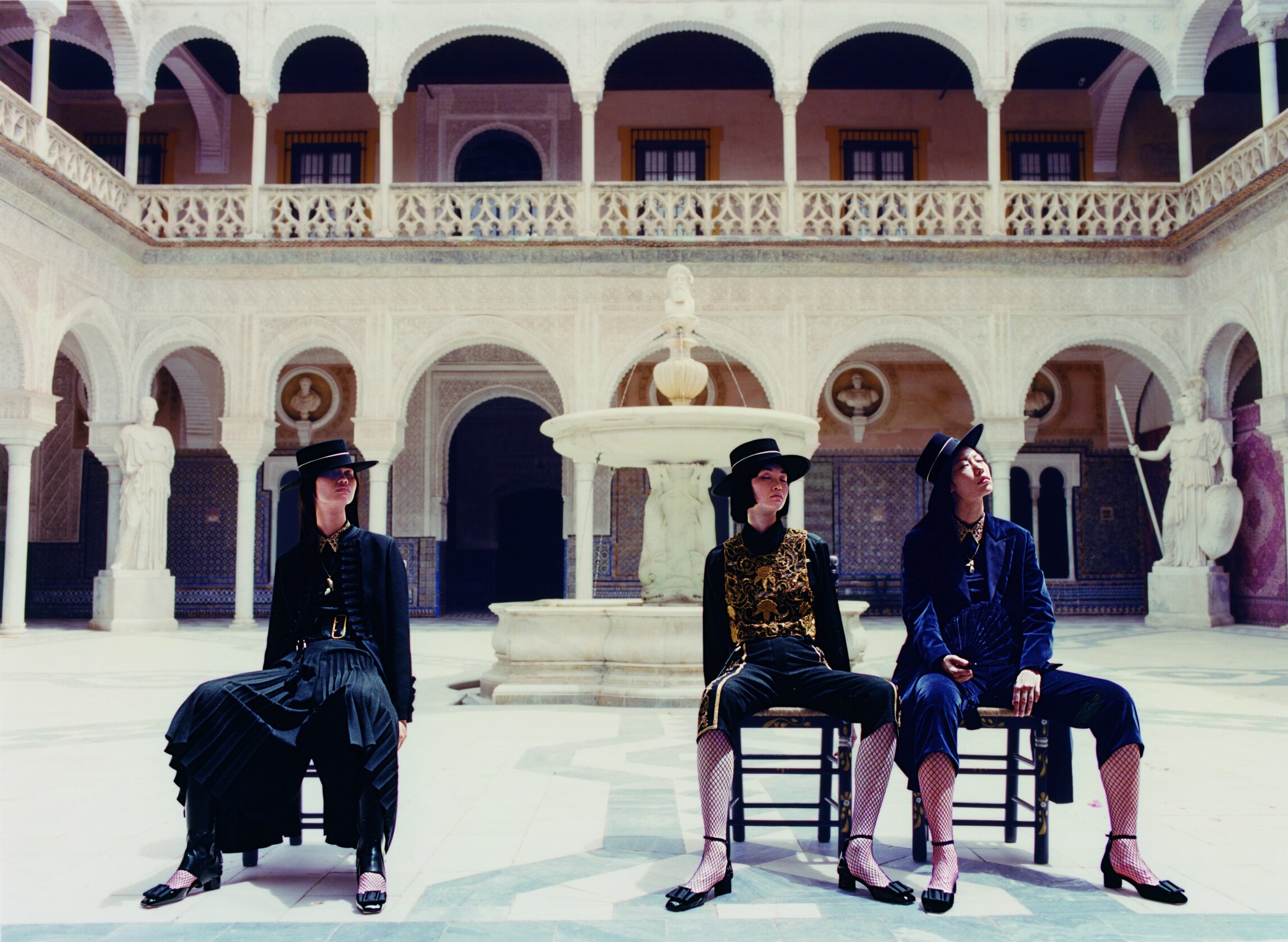 Three women seated in an elegant outdoor courtyard wearing black hats, dark jackets, pleated pants, and fishnet stockings in a fashion shoot.
