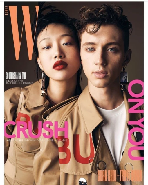 Fashion editorial magazine cover featuring an Asian woman with dark red lipstick and a Caucasian man in matching tan trench coats.