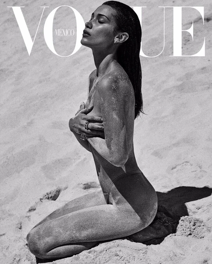 Monochrome fashion portrait of model Bella Hadid kneeling in sand on a beach with wet hair and sand dusted on her body