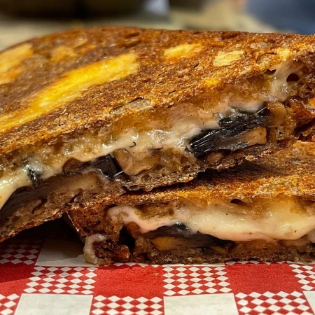 Grilled cheese sandwich with mushrooms cut in half on red and white checkered paper. Close-up of melting cheese and sliced mushrooms.