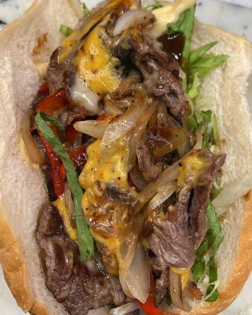 Delicious Philly cheesesteak sandwich on a white bun filled with sliced beef, melted cheese, grilled onions, peppers, and lettuce.
