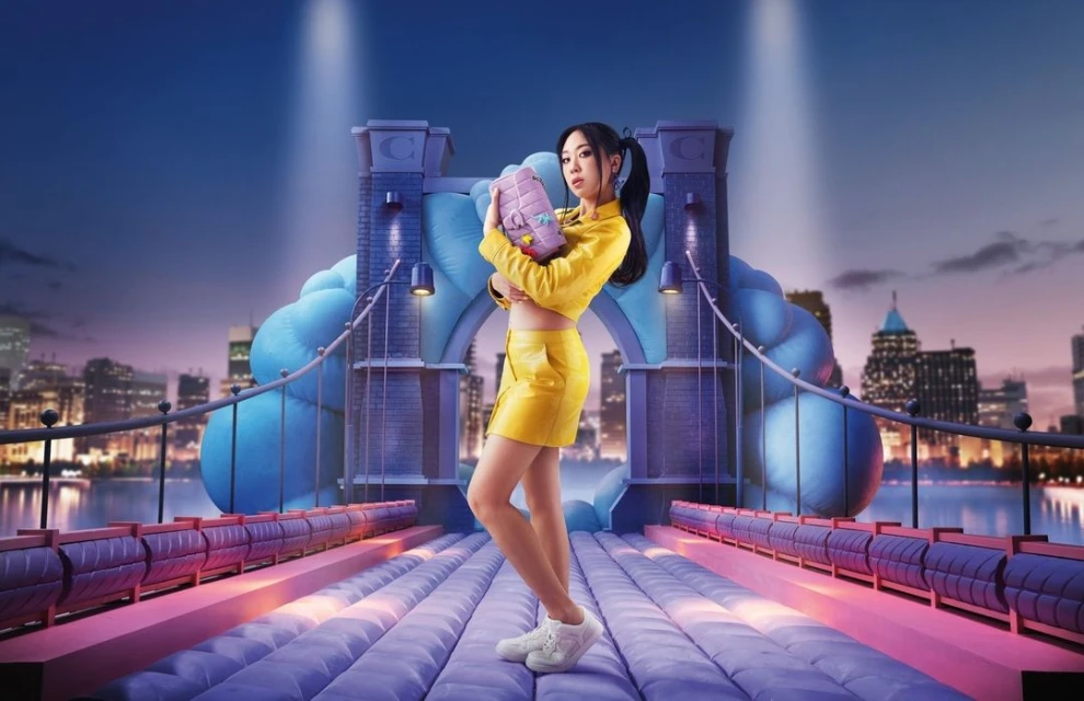 Woman in yellow outfit on a bridge with city skyline backdrop, holding a purple purse and wearing white sneakers, against a twilight sky.