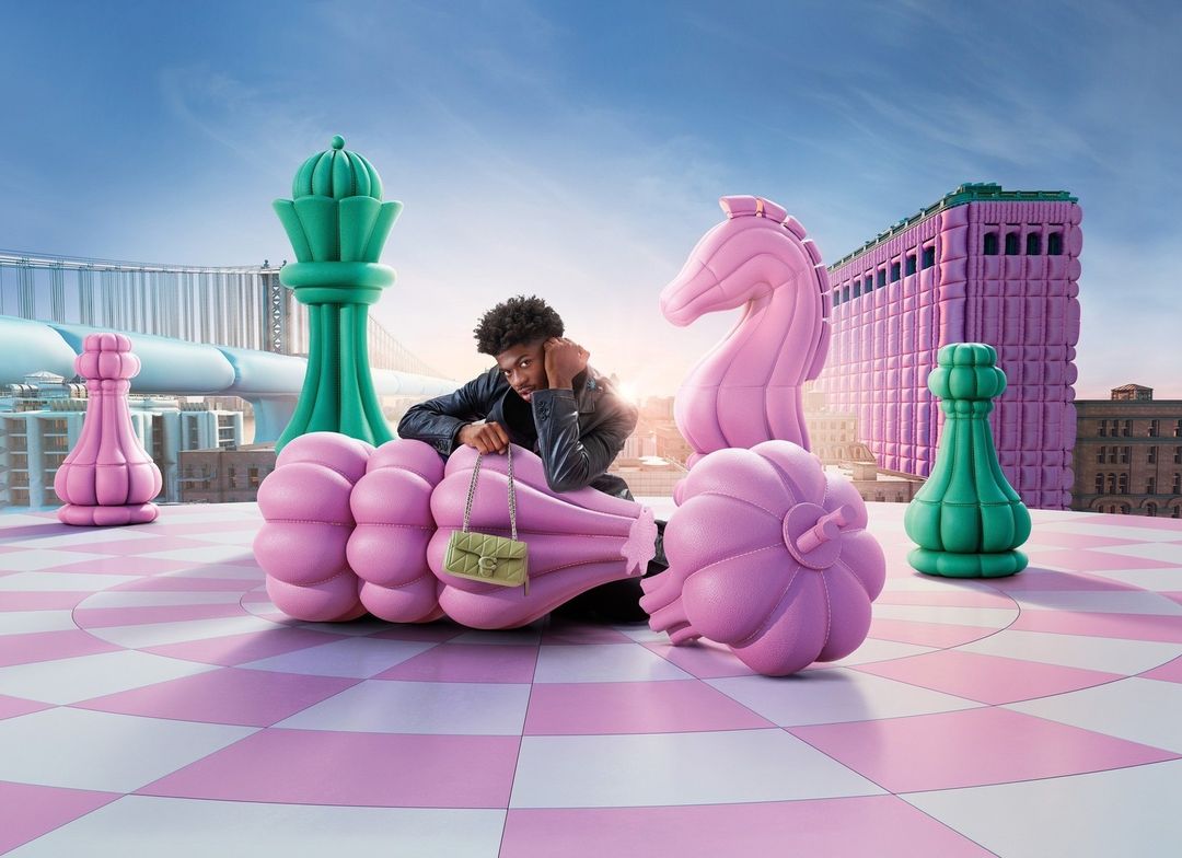 Man poses with a handbag and oversized chess pieces on a chessboard in front of a city skyline under a blue sky.