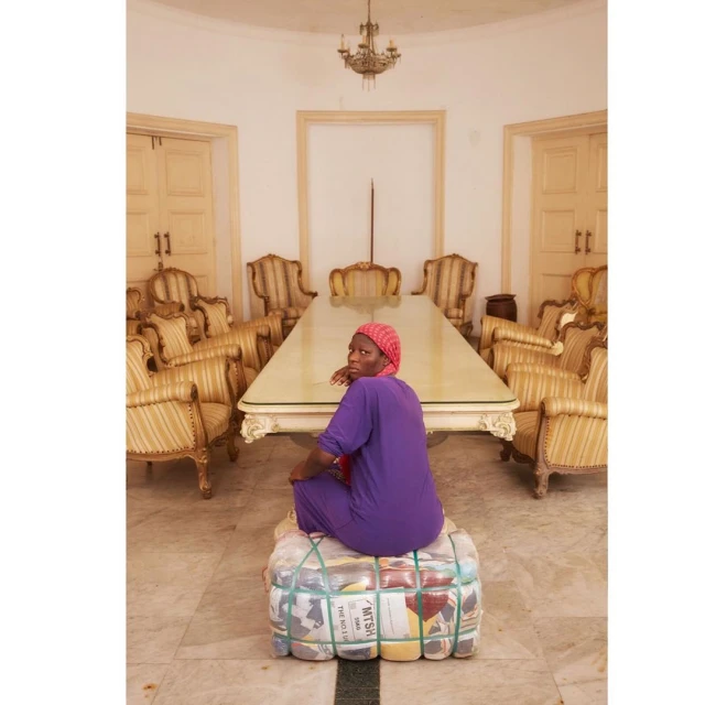 Woman in purple sits atop a fabric bale indoors, with formal chairs surrounding a table under a chandelier in a spacious room.