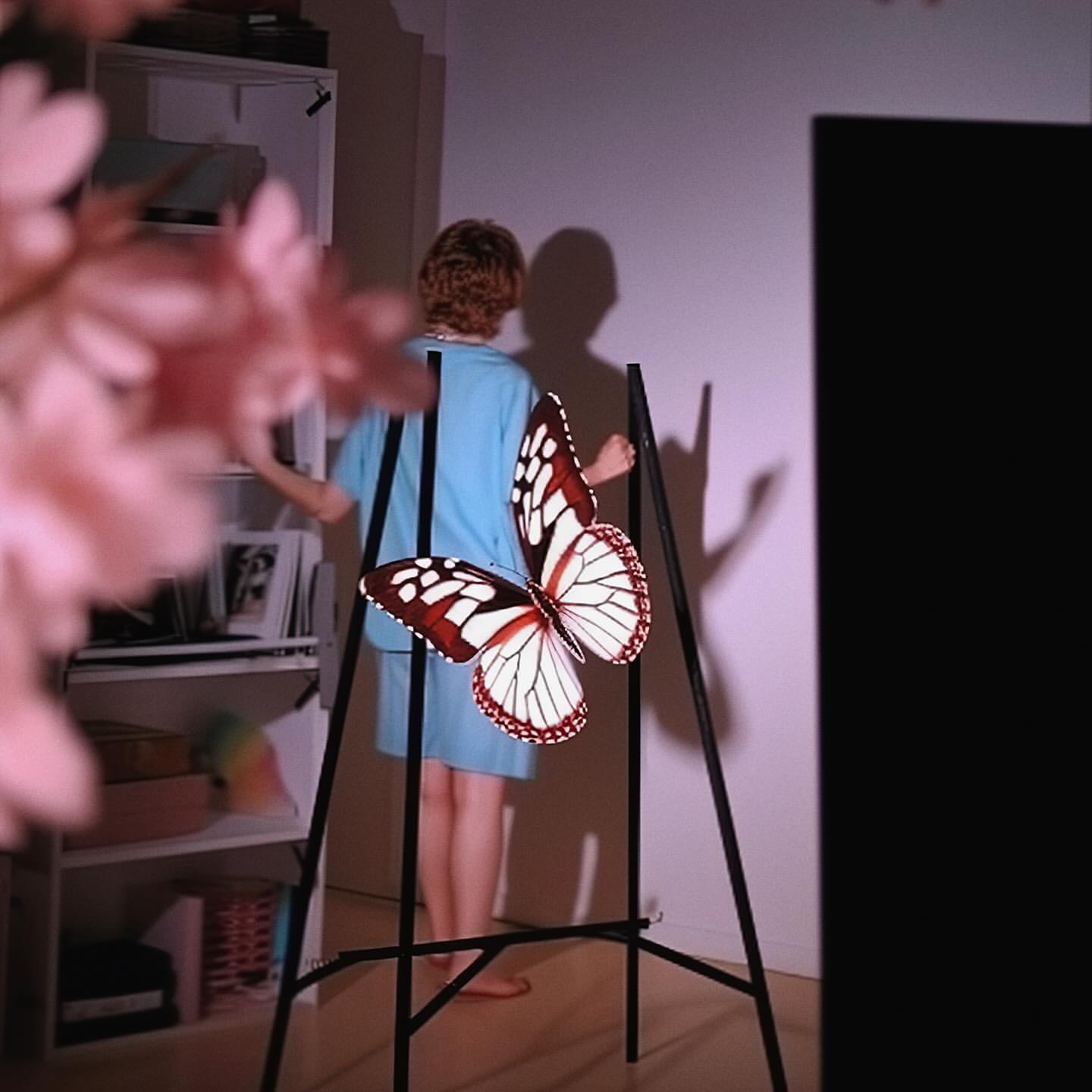 An artist tripod displaying a large butterfly art piece, a woman standing behind it indoors, with visible pink flowers and a shelf unit.