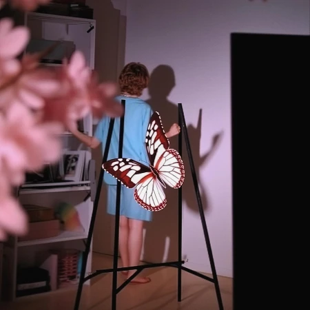 An artist tripod displaying a large butterfly art piece, a woman standing behind it indoors, with visible pink flowers and a shelf unit.