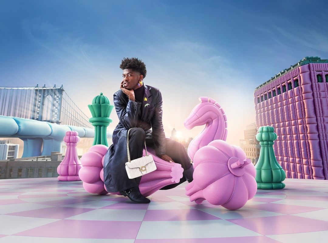 Lil Nas X poses on giant pink chess piece with white purse, wearing a black jacket and boots, with a city skyline and blue sky backdrop.