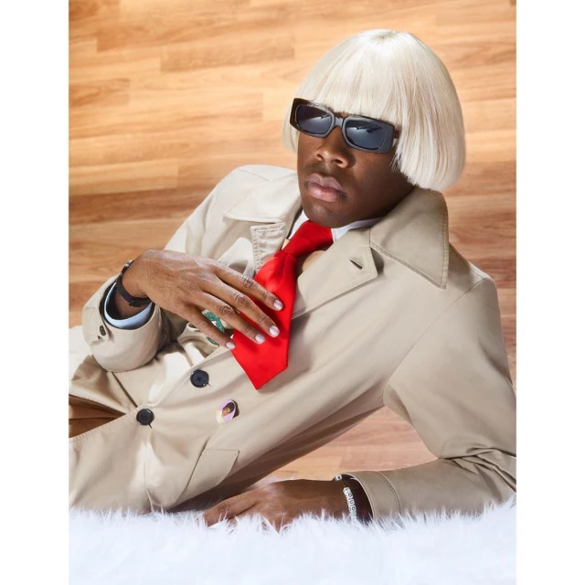 Man in beige trench coat and blonde wig poses with a red tie and sunglasses against a backdrop of wood grain and a white fur rug.