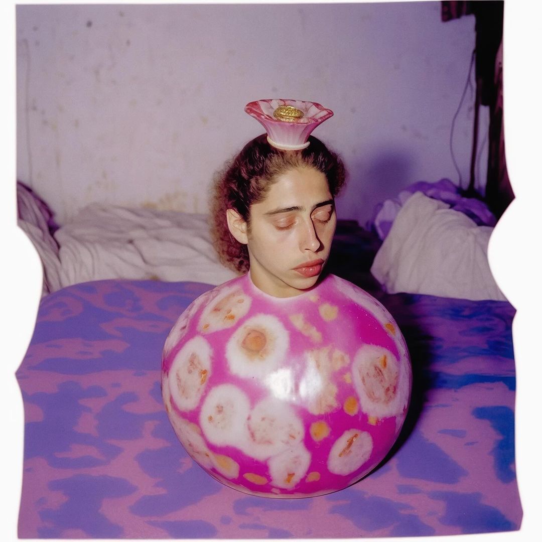 Young woman with closed eyes, wearing a pink and white spherical garment, with a pink flower-shaped hat indoors on a purple patterned bedspread.