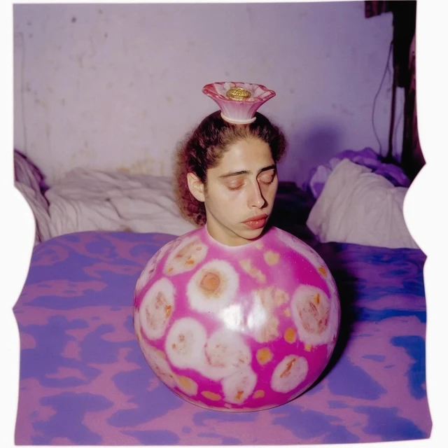 Young woman with closed eyes, wearing a pink and white spherical garment, with a pink flower-shaped hat indoors on a purple patterned bedspread.