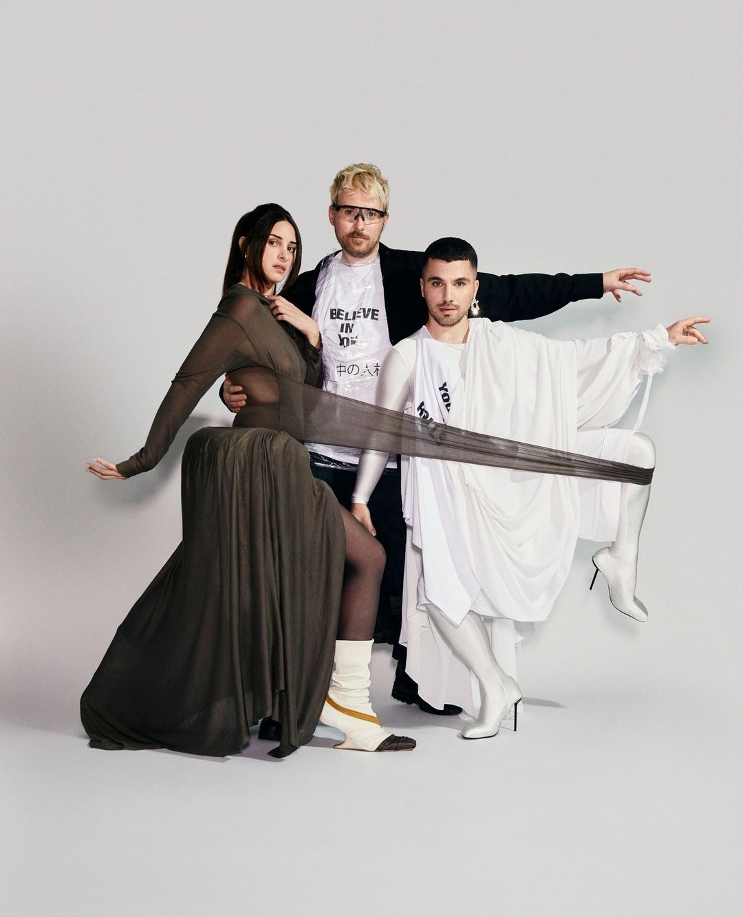 Three models in avant-garde fashion pose in a studio setting, featuring draped fabrics, statement boots, and unique accessories against a white backdrop.