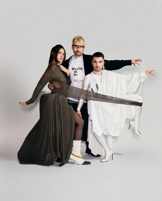 Three models in avant-garde fashion pose in a studio setting, featuring draped fabrics, statement boots, and unique accessories against a white backdrop.