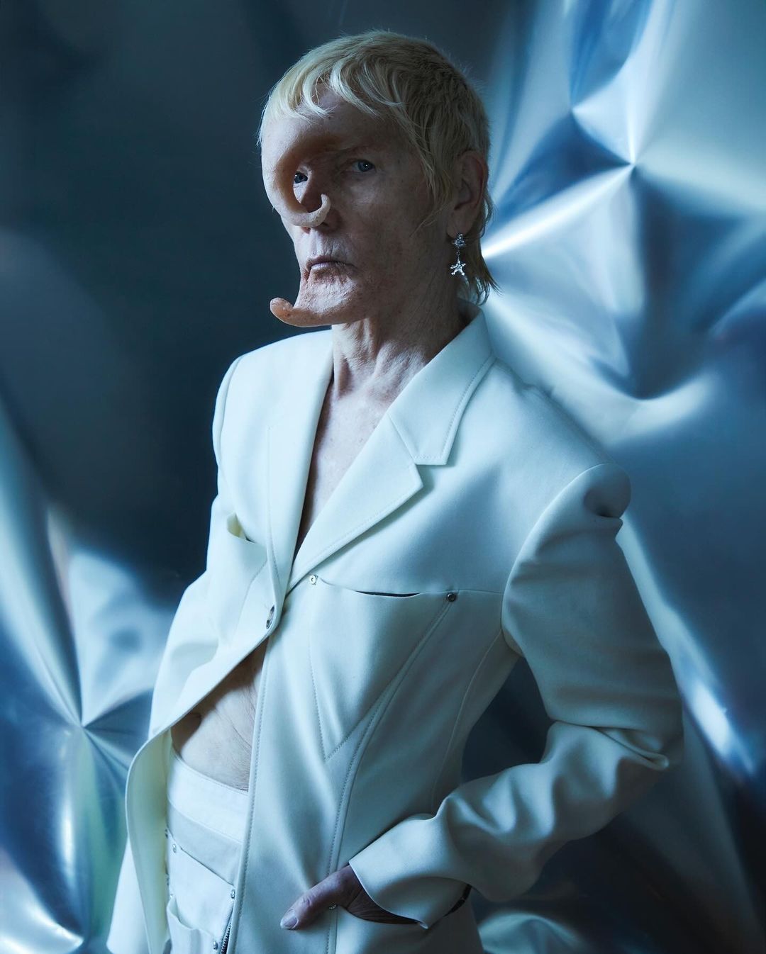 Unique individual in a white suit poses against a metallic background, highlighted by soft lighting and fashion-forward design.