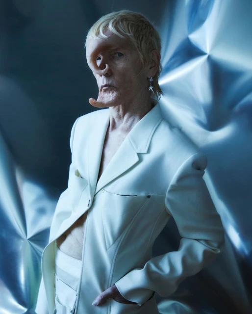 Unique individual in a white suit poses against a metallic background, highlighted by soft lighting and fashion-forward design.