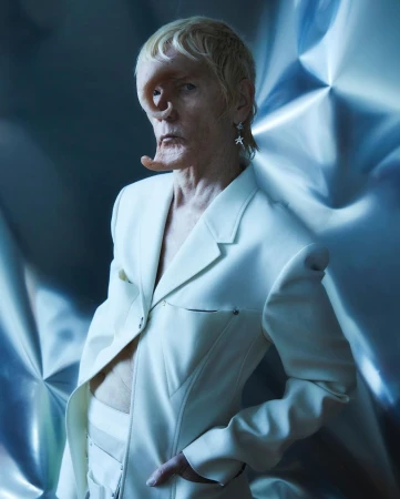 Unique individual in a white suit poses against a metallic background, highlighted by soft lighting and fashion-forward design.
