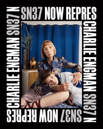 Older and younger adult embrace indoors. A blue curtain backdrop and surrounding advertisement text frame the subjects.