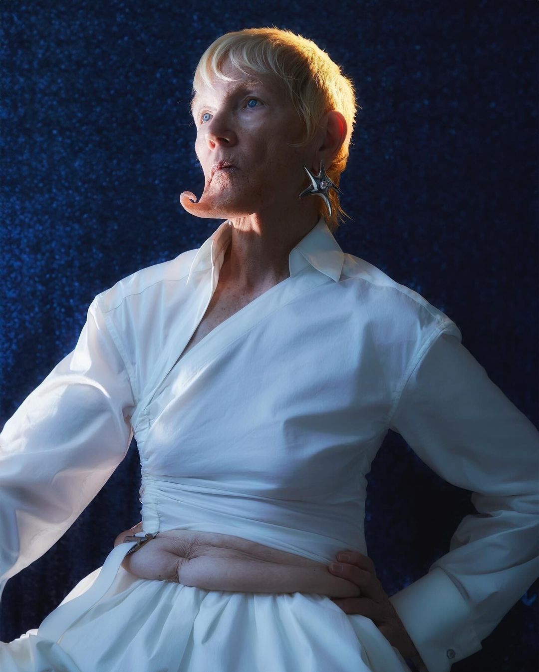 Portrait of a person with blonde hair wearing a white wrap shirt against a dark textured background
