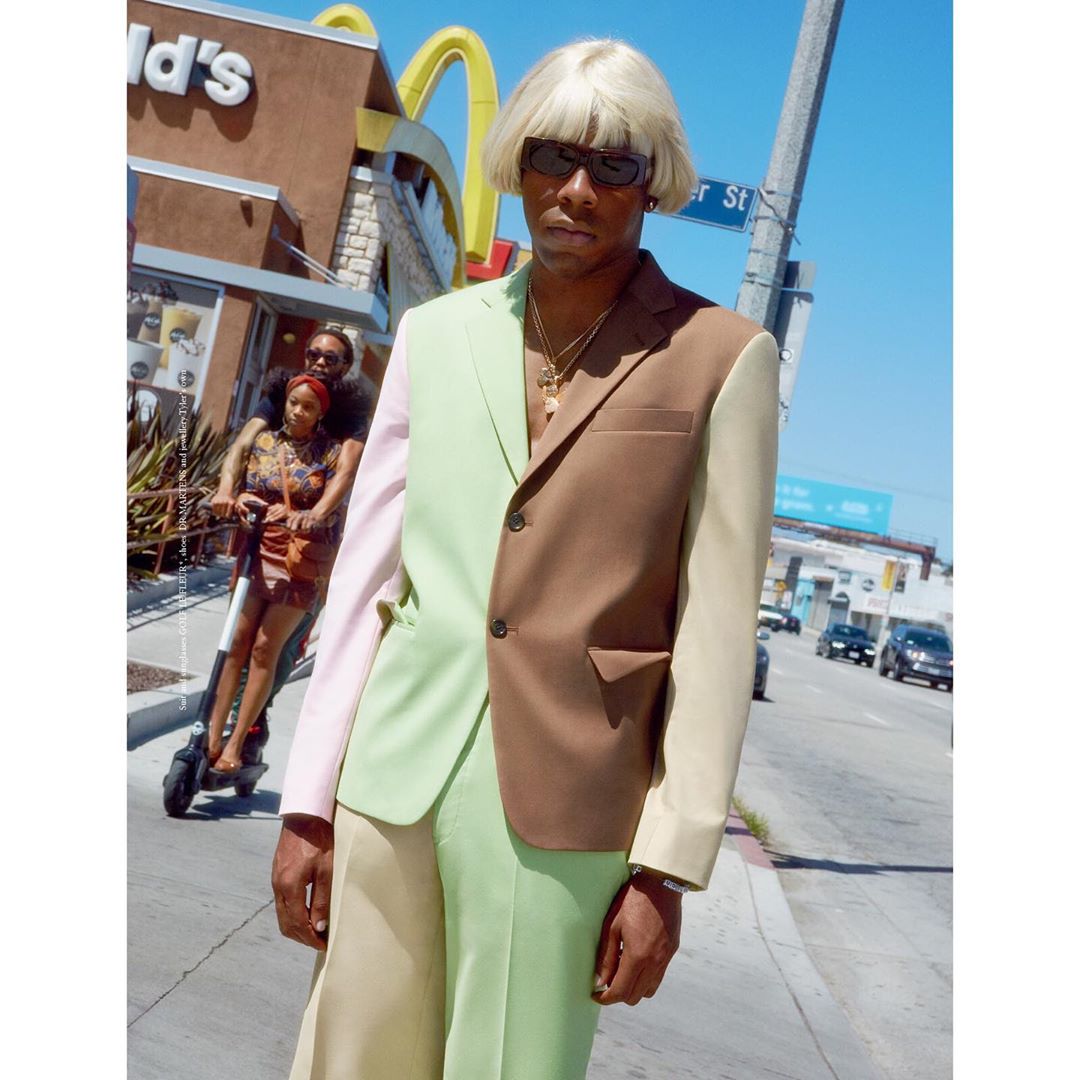Tyler, the Creator, poses in a color-blocked suit on a city street, with pedestrians on scooters and a McDonald's in the background.
