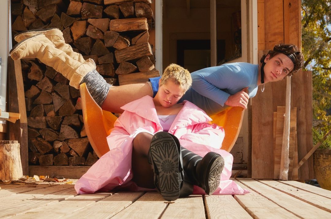 Two people on a wooden porch, one sleeping in a chair covered by a pink blanket, the other leaning over with a curious expression.