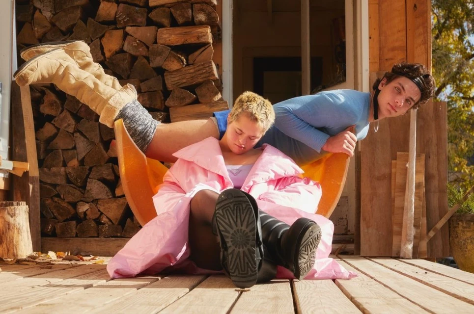 Two people on a wooden porch, one sleeping in a chair covered by a pink blanket, the other leaning over with a curious expression.