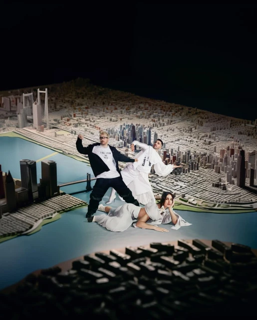 Three individuals pose with a scale model city featuring detailed buildings and a blue waterway, set against a dark background.
