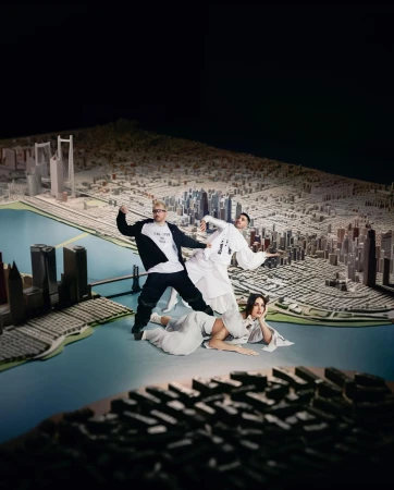 Three individuals pose with a scale model city featuring detailed buildings and a blue waterway, set against a dark background.