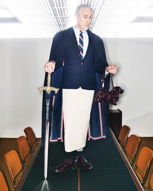 An adult male in formal wear stands on a conference table holding a dog purse and sword in an office with chairs.