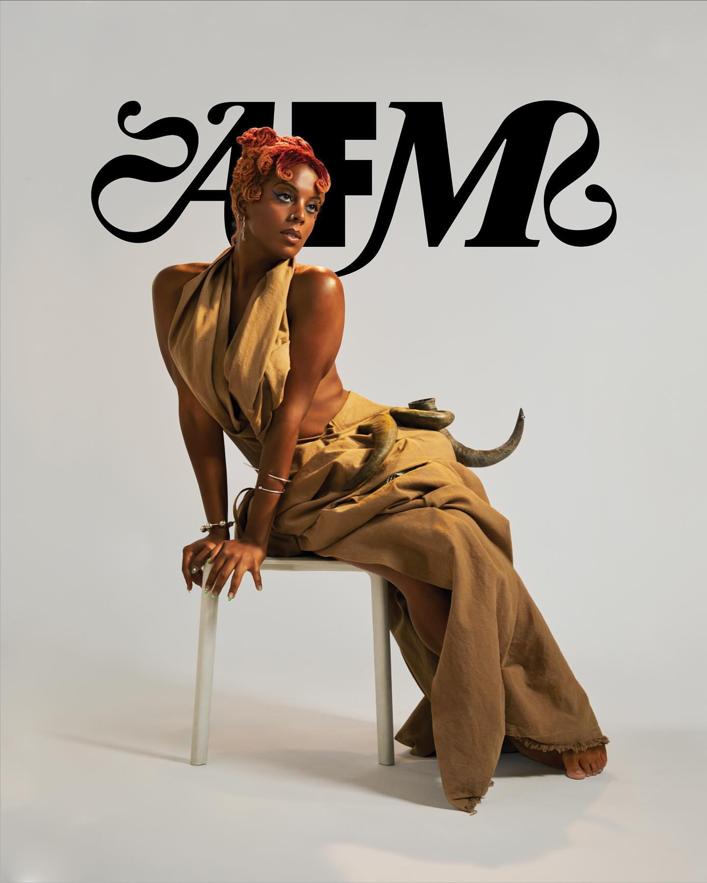 Elegant African-American woman in draped tan dress sitting on a chair against a neutral background with decorative animal object.