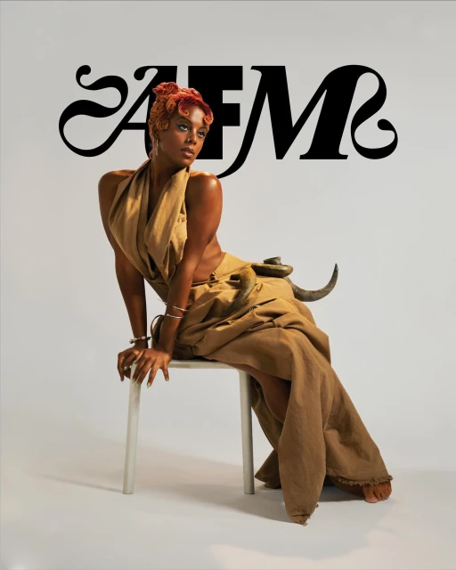 Elegant African-American woman in draped tan dress sitting on a chair against a neutral background with decorative animal object.