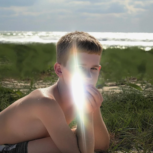 Young boy sits near the ocean with bright sunlight illuminating his face, set against a backdrop of sand and gentle waves.
