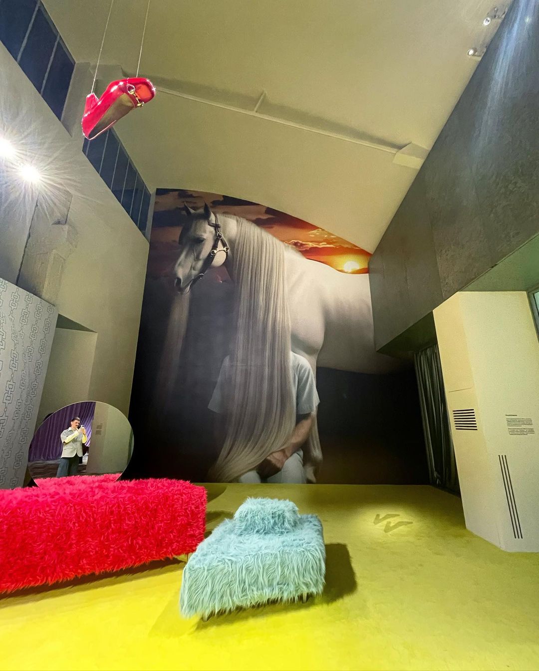 Art installation featuring a large horse mural, furry furniture, and abstract hanging shoe in a modern interior space with yellow flooring.