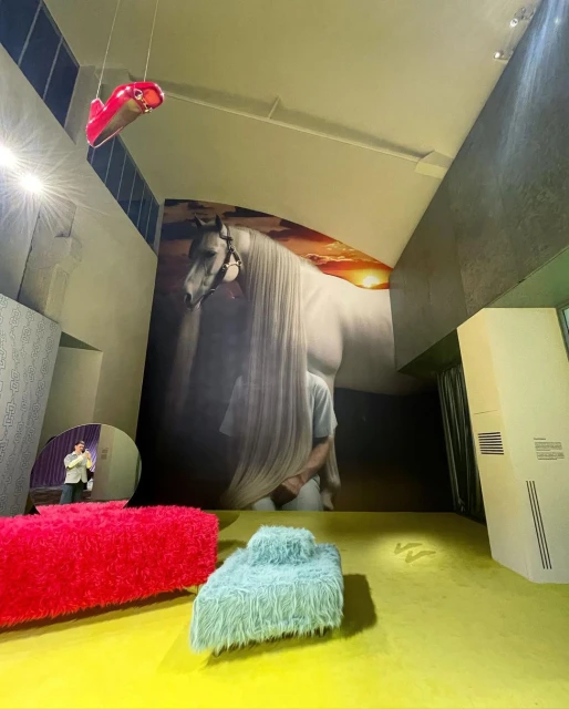 Art installation featuring a large horse mural, furry furniture, and abstract hanging shoe in a modern interior space with yellow flooring.