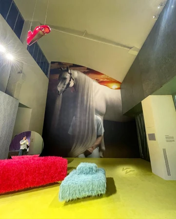 Art installation featuring a large horse mural, furry furniture, and abstract hanging shoe in a modern interior space with yellow flooring.