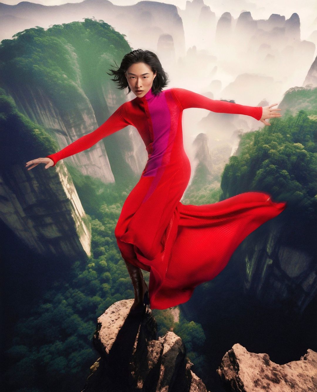 Woman in long red dress on rocky peak, arms outstretched, scenic mountain background. Fashion editorial, beauty, nature, adventure.