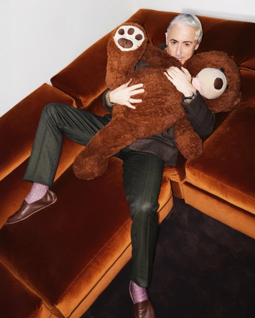 Adult male lying on a rust-colored sectional sofa, hugging a large brown teddy bear, wearing brown shoes and green trousers.