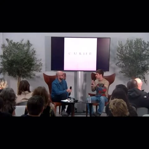 Two people are seated on stage for a discussion in front of an audience. A screen displays the text 'CURSED'. Indoor event setting.