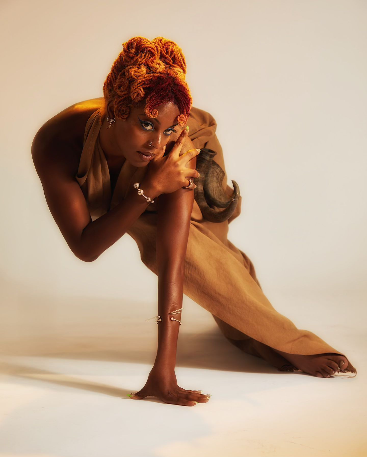 A dark-skinned adult female in a tan jumpsuit poses in a low stance with orange-red hair on a seamless gradient beige background.
