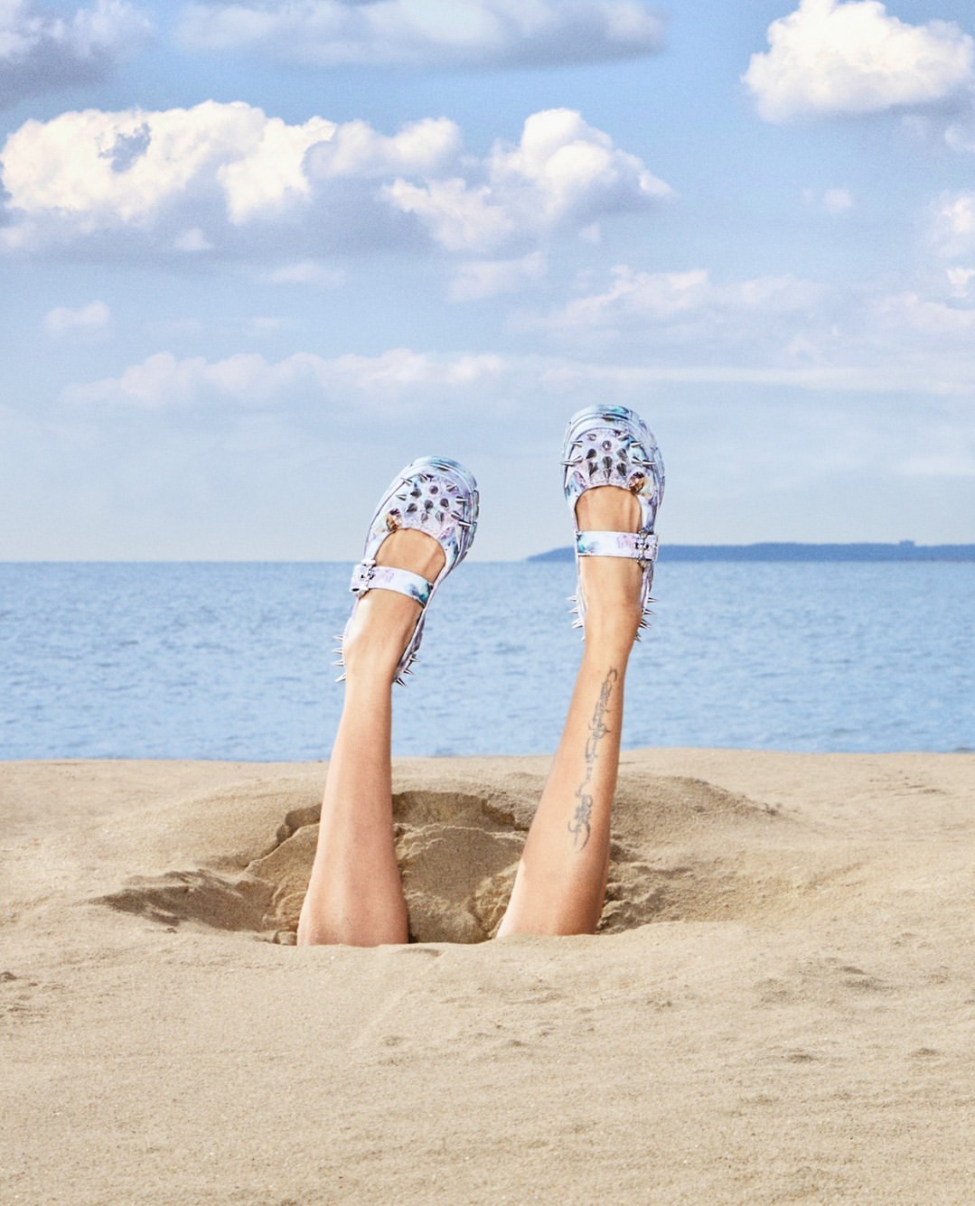 Legs buried in sand wearing patterned strappy shoes against an ocean and sky background