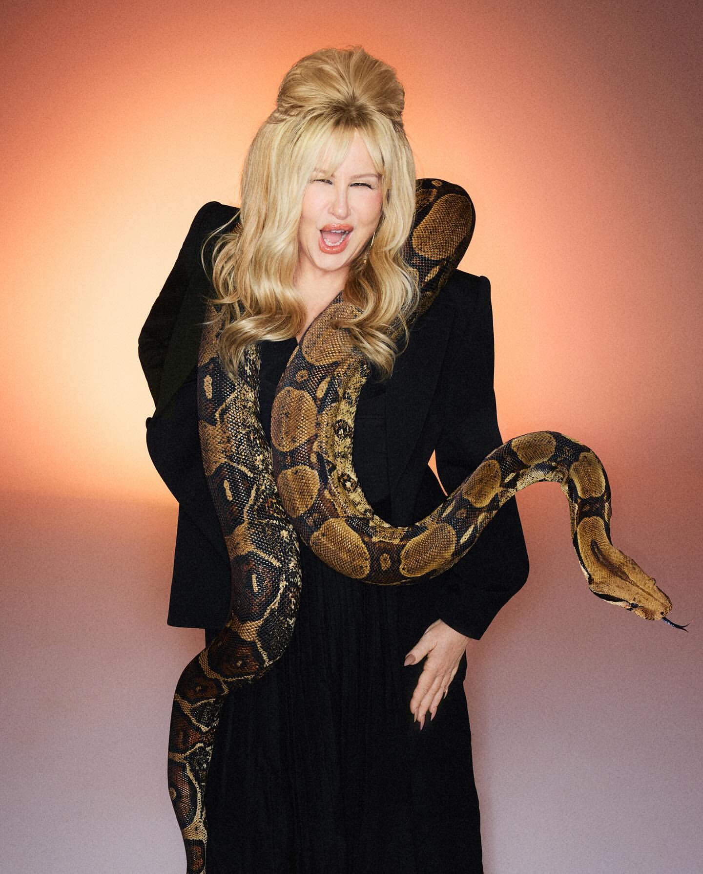 Jennifer Coolidge poses in a studio with a large python draped around her shoulders, wearing an elegant black jacket and dress.