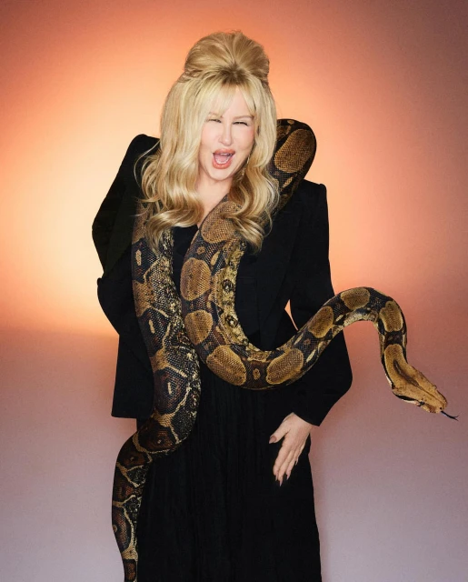 Jennifer Coolidge poses in a studio with a large python draped around her shoulders, wearing an elegant black jacket and dress.