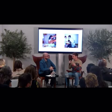 Two men sit on chairs, one speaking, in a room with an audience and large display showing medical images, trees in the background.
