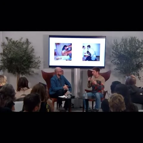 Two men sit on chairs, one speaking, in a room with an audience and large display showing medical images, trees in the background.