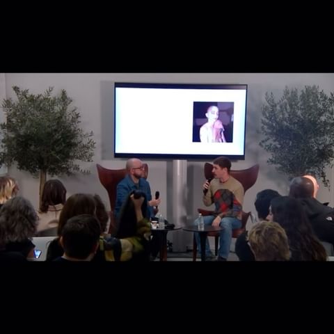 Two adult men sit on stage in chairs having a discussion in front of a seated audience, in an indoor setting with trees.