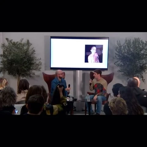 Two adult men sit on stage in chairs having a discussion in front of a seated audience, in an indoor setting with trees.