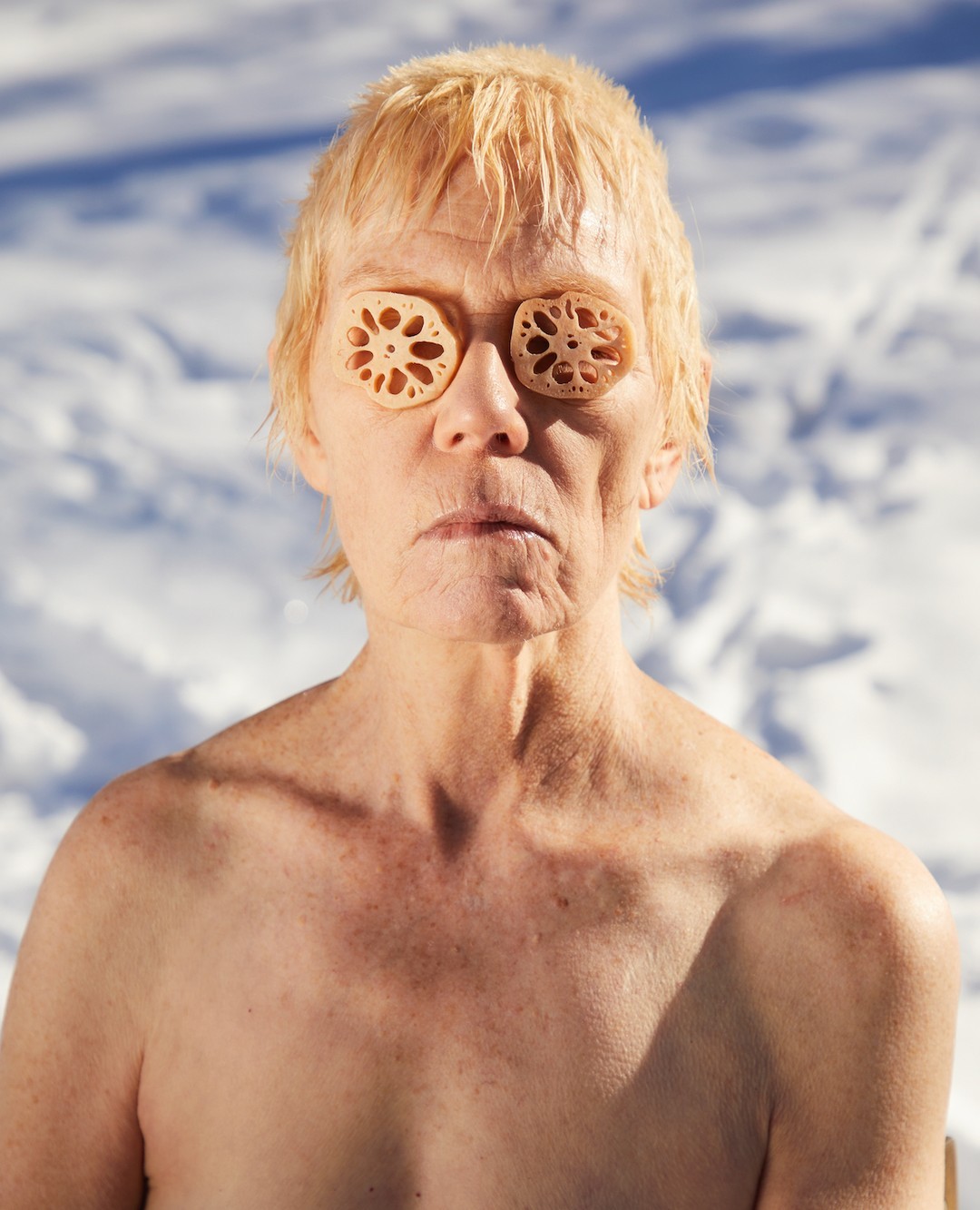 Middle-aged person with pale skin and blonde hair stands outdoors in winter snow, with two cross-sections of lotus root covering the eyes.