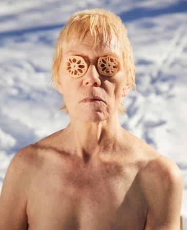 Middle-aged person with pale skin and blonde hair stands outdoors in winter snow, with two cross-sections of lotus root covering the eyes.