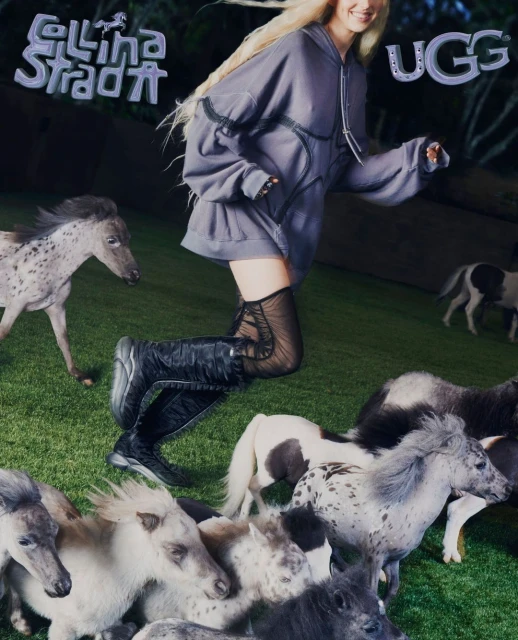 A woman in a grey hoodie and black boots runs across green grass, surrounded by a herd of small ponies in an outdoor setting.