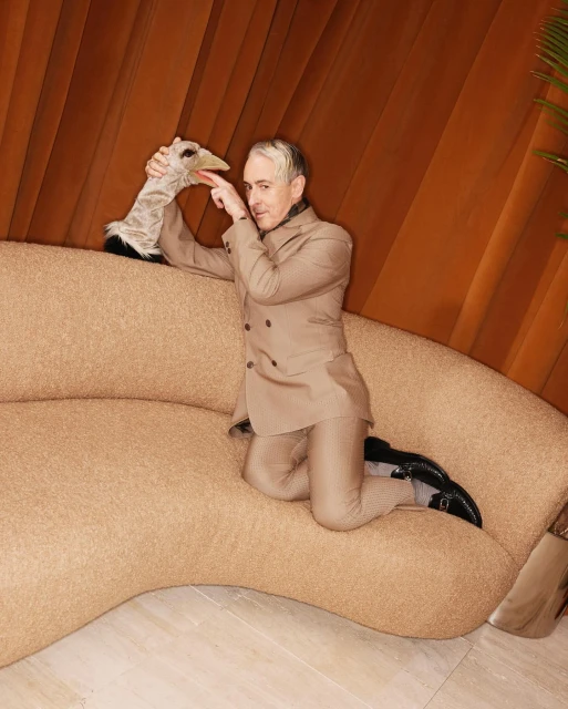 Man in a suit kneels on a light brown couch, touching the beak of an ostrich hand puppet, in front of an orange curtain.